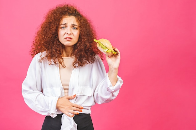 Mulher jovem com dor de estômago segurando um hambúrguer