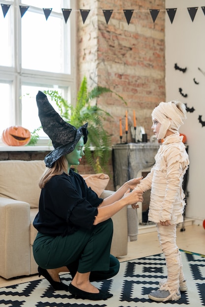 Mulher jovem com fantasia de bruxa agachada perto da poltrona e ajustando bandagens na fantasia de meninos em quarto decorado no Halloween