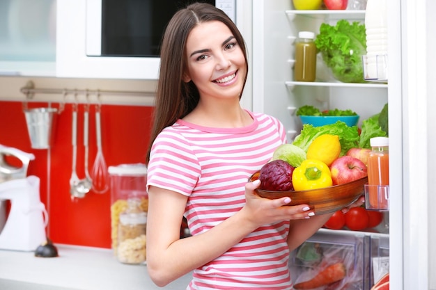 Mulher jovem com frutas e legumes ao lado da geladeira na cozinha