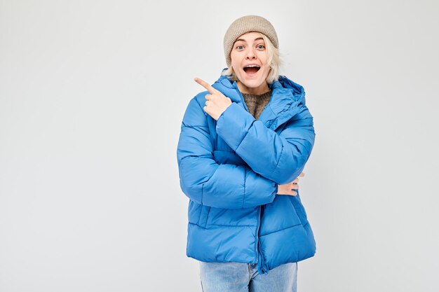 Foto mulher jovem com jaqueta azul de inverno apontando dedo para o lado demonstrando espaço vazio para produto ou texto isolado em fundo branco de estúdio