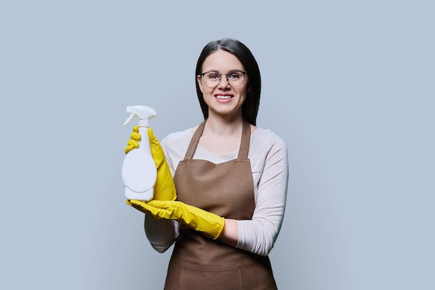 Foto mulher jovem com luvas avental com detergente orgânico nas mãos olhando para a câmera em fundo cinza claro serviço de limpeza doméstica limpeza natural eco detergentes orgânicos conceito de ecologia