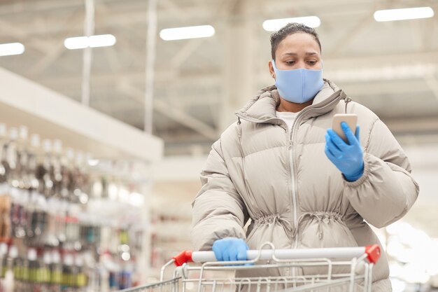 Mulher jovem com luvas de proteção e máscara lendo uma mensagem em seu telefone celular enquanto faz compras com o carrinho no supermercado