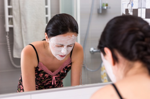 Mulher jovem com máscara cosmética lavando o rosto em frente ao espelho no banheiro moderno de azulejos de casa