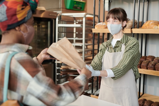 Mulher jovem com máscara dando pão fresco em pacote para o cliente na padaria