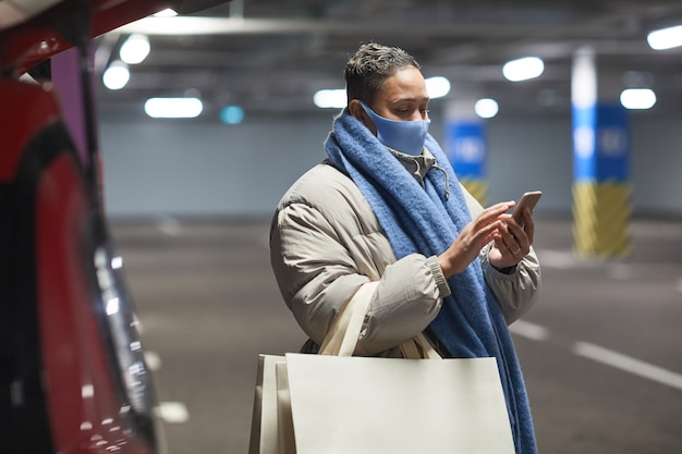 Mulher jovem com máscara protetora com sacolas de compras, usando telefone celular em um estacionamento subterrâneo