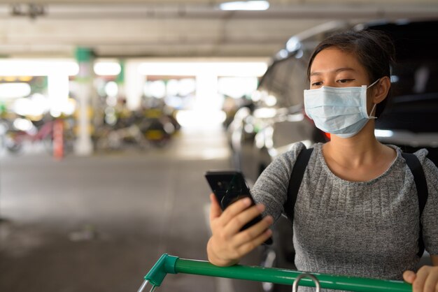 Mulher jovem com máscara usando telefone, segurando o carrinho de compras no estacionamento