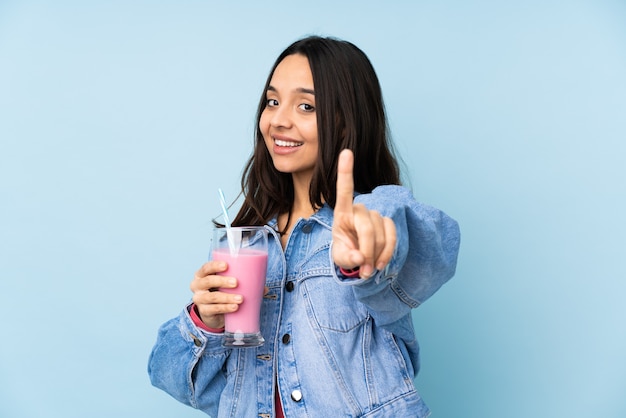 Mulher jovem com milk-shake de morango em azul isolado mostrando e levantando um dedo