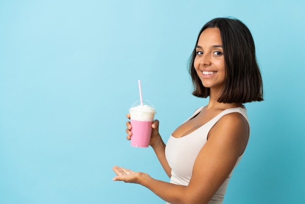 Mulher jovem com milk-shake de morango isolada na parede azul, estendendo as mãos para o lado para convidar para vir