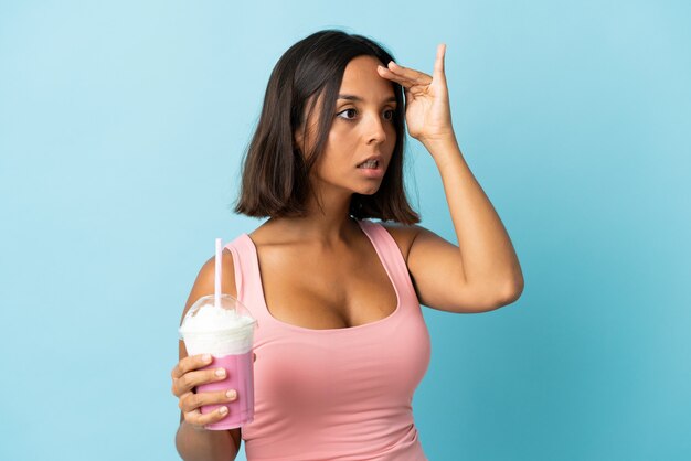 Mulher jovem com milk-shake de morango isolado na parede azul com expressão de surpresa enquanto olha para o lado