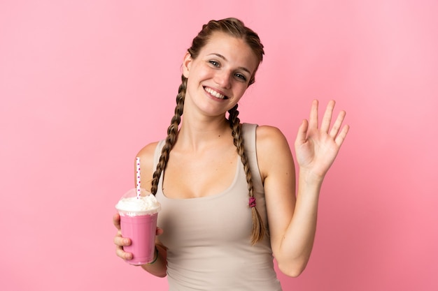 Mulher jovem com milk-shake de morango isolado na parede rosa saudando com a mão com expressão feliz