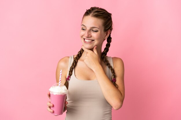 Mulher jovem com milkshake de morango isolada em um fundo rosa olhando para o lado e sorrindo