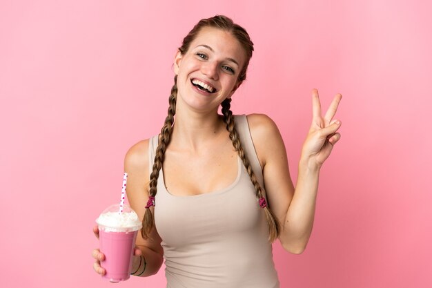 Mulher jovem com milkshake de morango isolada em um fundo rosa sorrindo e mostrando sinal de vitória