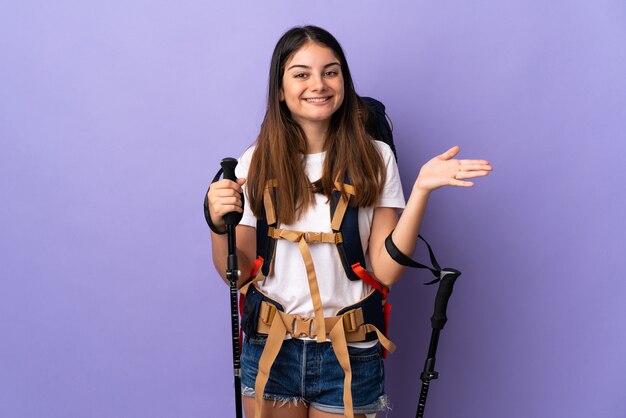 Mulher jovem com mochila e bastões de trekking isolados em um roxo estendendo as mãos para o lado para convidar para vir