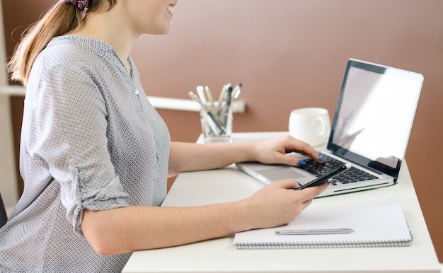 Mulher jovem com óculos, trabalhando em casa usando telefone inteligente e computador, mãos de mulher usando telefone inteligente