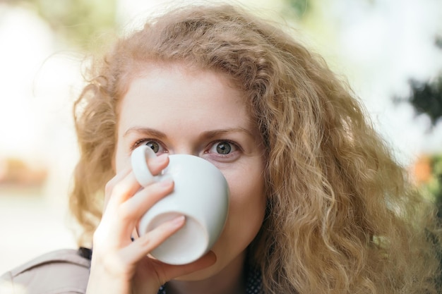 Mulher jovem com rosto bonito e cabelo comprido encaracolado bebendo café branco ou xícara de chá ao ar livre