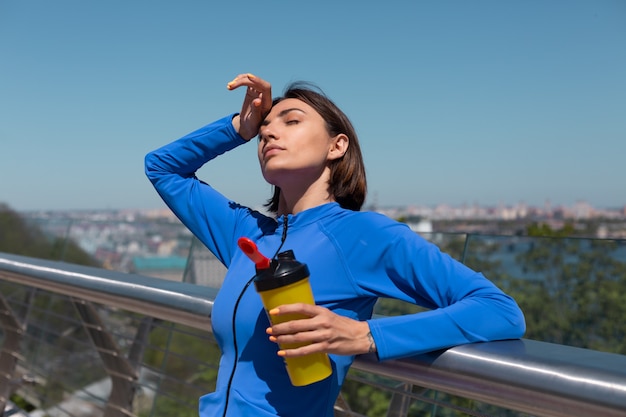 Mulher jovem com roupa esportiva adequada na ponte em uma manhã quente e ensolarada com uma garrafa de shaker com sede após o treino, cansada de beber