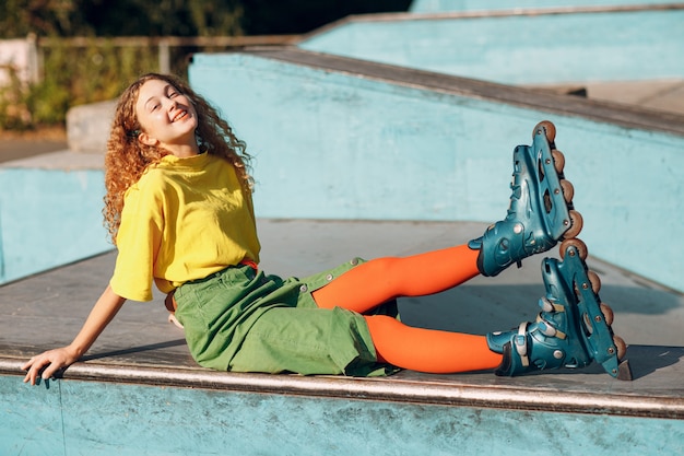 Mulher jovem com roupas verdes e amarelas e meias laranja com penteado encaracolado patinando na pista de skate
