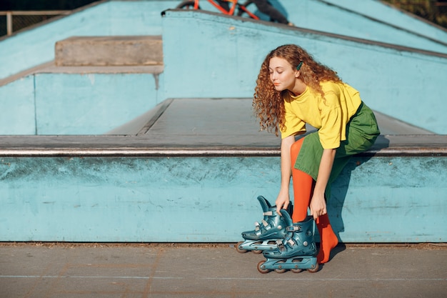 Mulher jovem com roupas verdes e amarelas e meias laranja com penteado encaracolado patinando na pista de skate