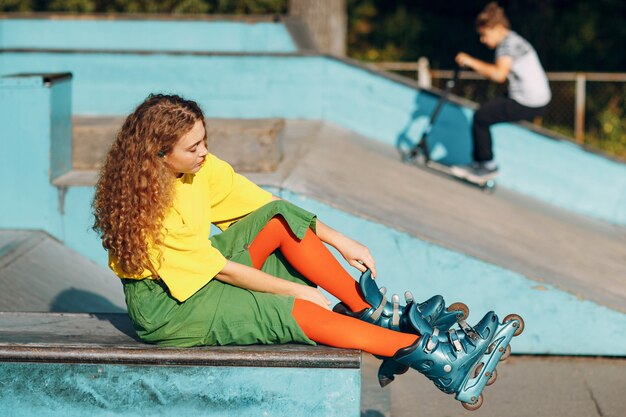 Mulher jovem com roupas verdes e amarelas e meias laranja com penteado encaracolado patinando no parque de skate