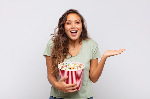 Mulher jovem com um balde de pop se sentindo feliz, animada, surpresa ou chocada, sorrindo e surpresa com algo inacreditável