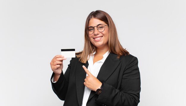 Mulher jovem com um cartão de crédito sorrindo alegremente, sentindo-se feliz e apontando para o lado e para cima, mostrando o objeto no espaço da cópia