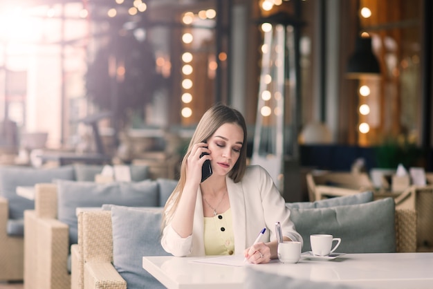 Mulher jovem com um casaco branco a trabalhar num café com jornais, a beber café
