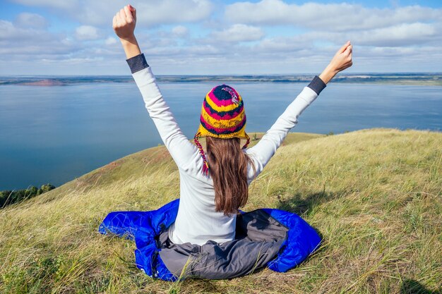 Mulher jovem com um chapéu engraçado do nepal em um saco de dormir azul quente, fazendo o aquecimento da manhã, alongando-se contra o fundo de uma bela paisagem de lago