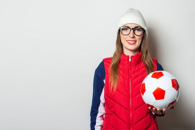 Mulher jovem com um colete vermelho, chapéu e óculos segurando uma bola de futebol contra uma parede de luz