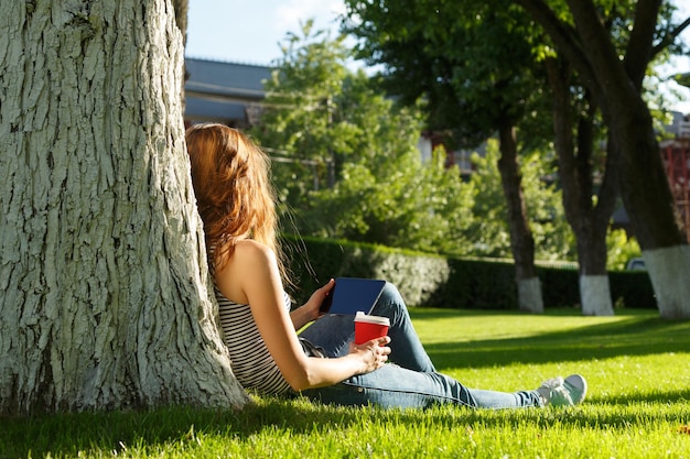 Mulher jovem com um copo de papel descartável vermelho de café usando o tablet pc sob a árvore em um gramado verde em um parque