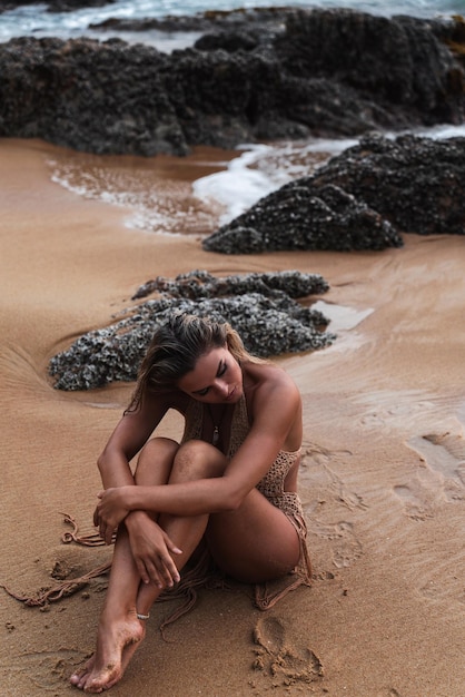 Mulher jovem com um corpo bronzeado impecável e bonita maquiagem vestindo um vestido de crochet chique enquanto ela senta-se graciosamente na praia