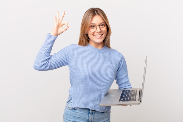 Mulher jovem com um laptop se sentindo feliz, mostrando aprovação com um gesto de ok