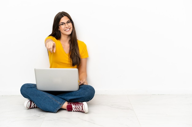 Mulher jovem com um laptop sentada no chão aponta o dedo para você com uma expressão confiante