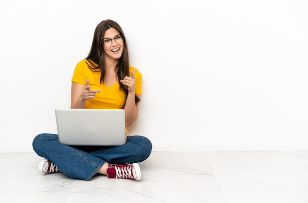 Mulher jovem com um laptop sentada no chão, apontando para a frente e sorrindo