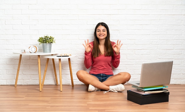 Mulher jovem com um laptop sentada no chão dentro de casa, mostrando sinal de ok com as duas mãos