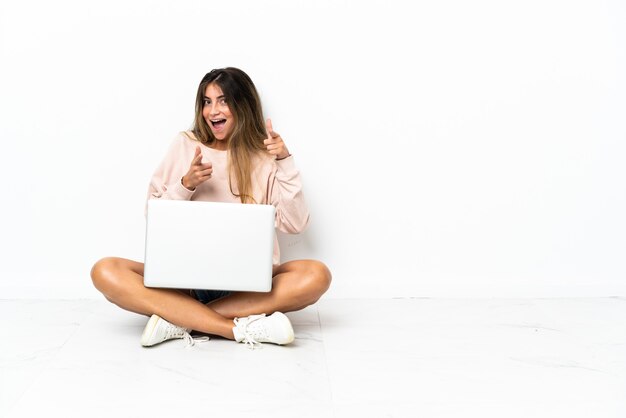 Mulher jovem com um laptop sentada no chão, isolada na parede branca, apontando para a frente e sorrindo