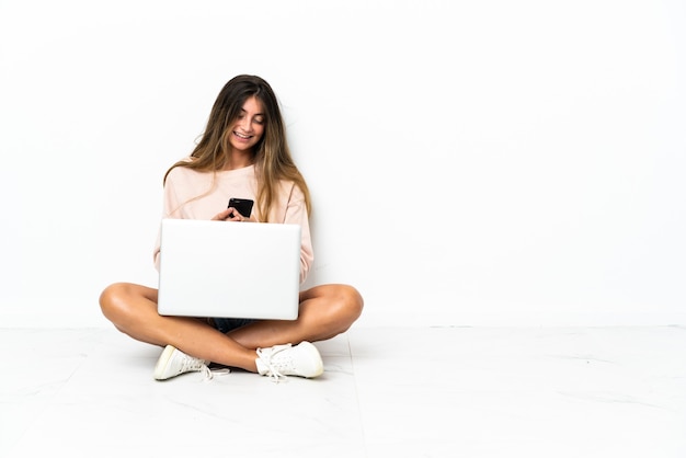 Mulher jovem com um laptop sentada no chão, isolada na parede branca, enviando uma mensagem com o celular