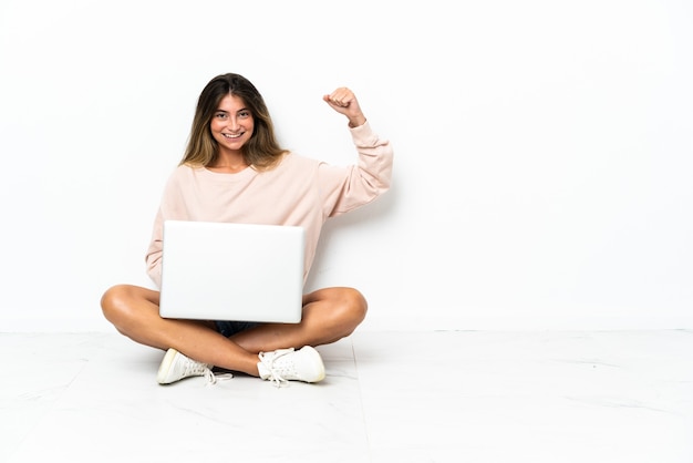 Mulher jovem com um laptop sentada no chão, isolada na parede branca, fazendo um gesto forte