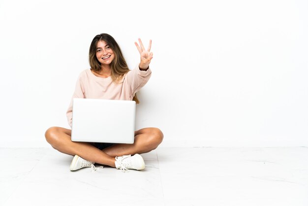 Mulher jovem com um laptop sentada no chão, isolada na parede branca, feliz e contando três com os dedos