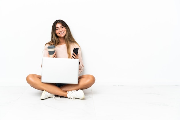 Mulher jovem com um laptop sentada no chão, isolada na parede branca, segurando um café para levar e um celular