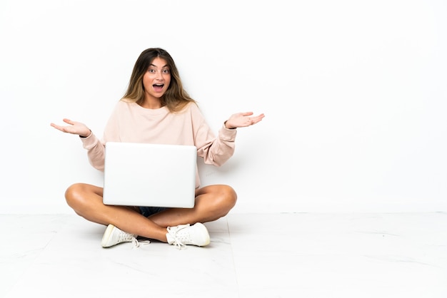 Mulher jovem com um laptop sentada no chão, isolada no fundo branco, com expressão facial chocada