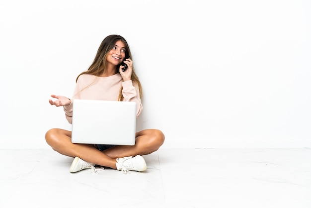 Mulher jovem com um laptop sentada no chão, isolada no fundo branco, conversando com alguém ao telefone celular