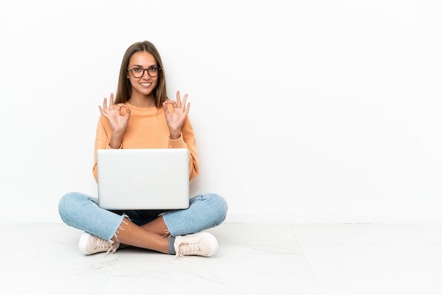 Mulher jovem com um laptop sentada no chão, mostrando um sinal de ok com os dedos