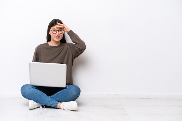 Mulher jovem com um laptop sentada no chão olhando para longe com a mão para olhar algo