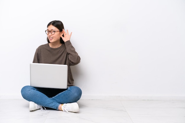 Mulher jovem com um laptop sentada no chão ouvindo algo colocando a mão na orelha