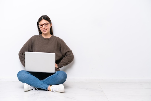 Mulher jovem com um laptop sentada no chão, posando com os braços na cintura e sorrindo