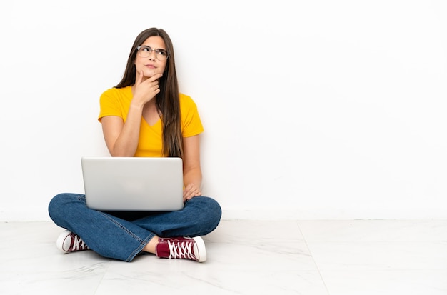 Mulher jovem com um laptop sentada no chão, tendo dúvidas e com uma expressão facial confusa