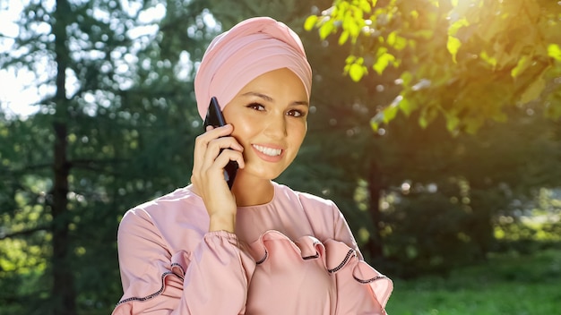 Mulher jovem com um lindo turbante e um vestido da mesma cor rosa suave está falando ao telefone contra o fundo de árvores