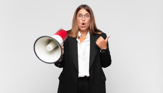 Mulher jovem com um megafone parecendo surpresa e descrente, apontando para um objeto ao lado e dizendo uau, inacreditável