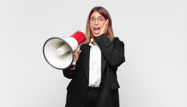 Mulher jovem com um megafone se sentindo feliz, animada e surpresa, olhando para o lado com as duas mãos no rosto