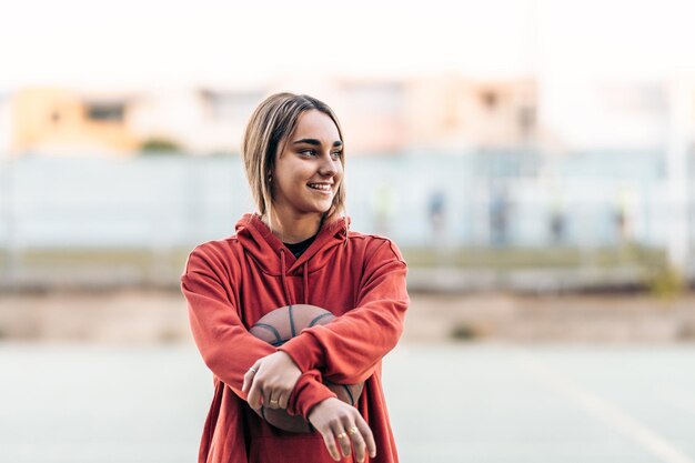 Mulher jovem com um moletom esportivo olhando para o lado enquanto segura uma bola de basquete em uma quadra ao ar livre